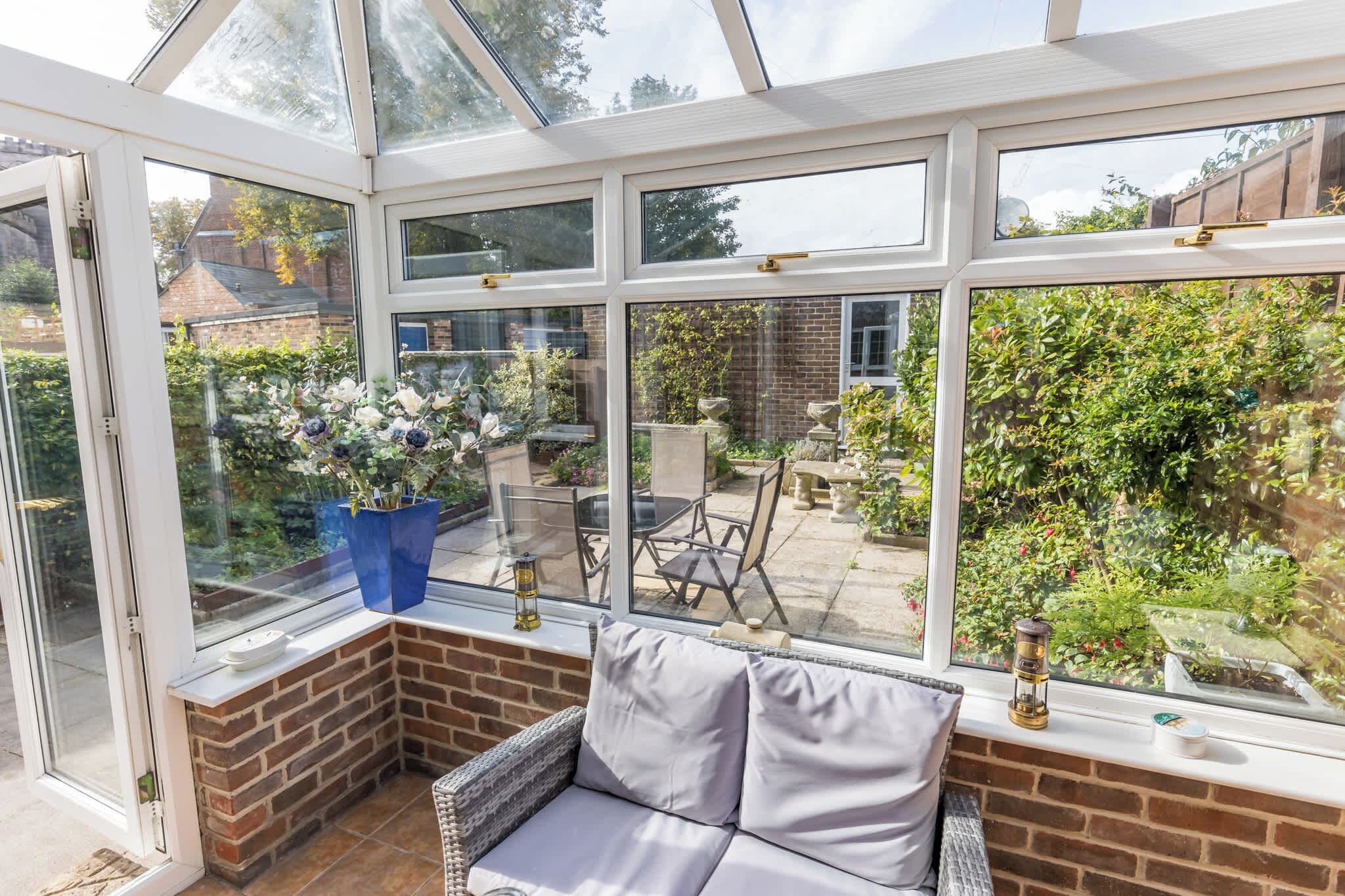 A conservatory with a glass roof, a wicker sofa, and a view of a garden with plants and a patio area.