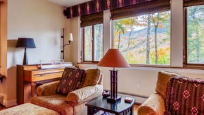 A cozy living room with a wooden desk, two armchairs, a table with a lamp, and large windows offering a view of trees and hills. - Gavia Chalet - Mountain Views - Private Hot Tub - Loon Mtn - Kancamangus Highway (Lincoln)