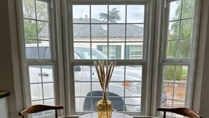 A corner room with a bay window, a round table with chairs, and a vase with reed sticks.