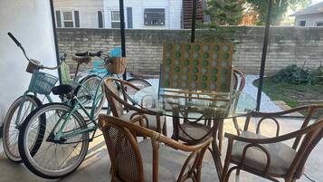 Un patio con sillas de mimbre, una mesa de vidrio, una bicicleta y vistas a una calle residencial.