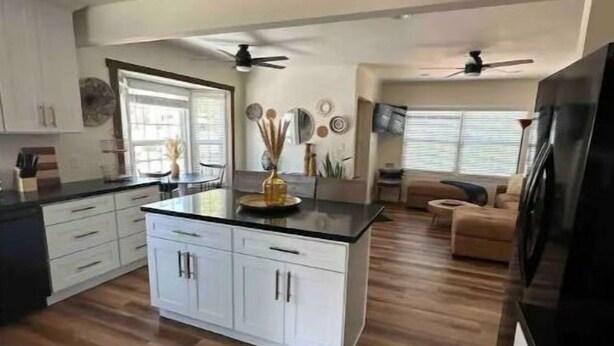 A spacious kitchen with white cabinets, a black countertop, and a central island. There are ceiling fans, a large window, and a television mounted on the wall.
