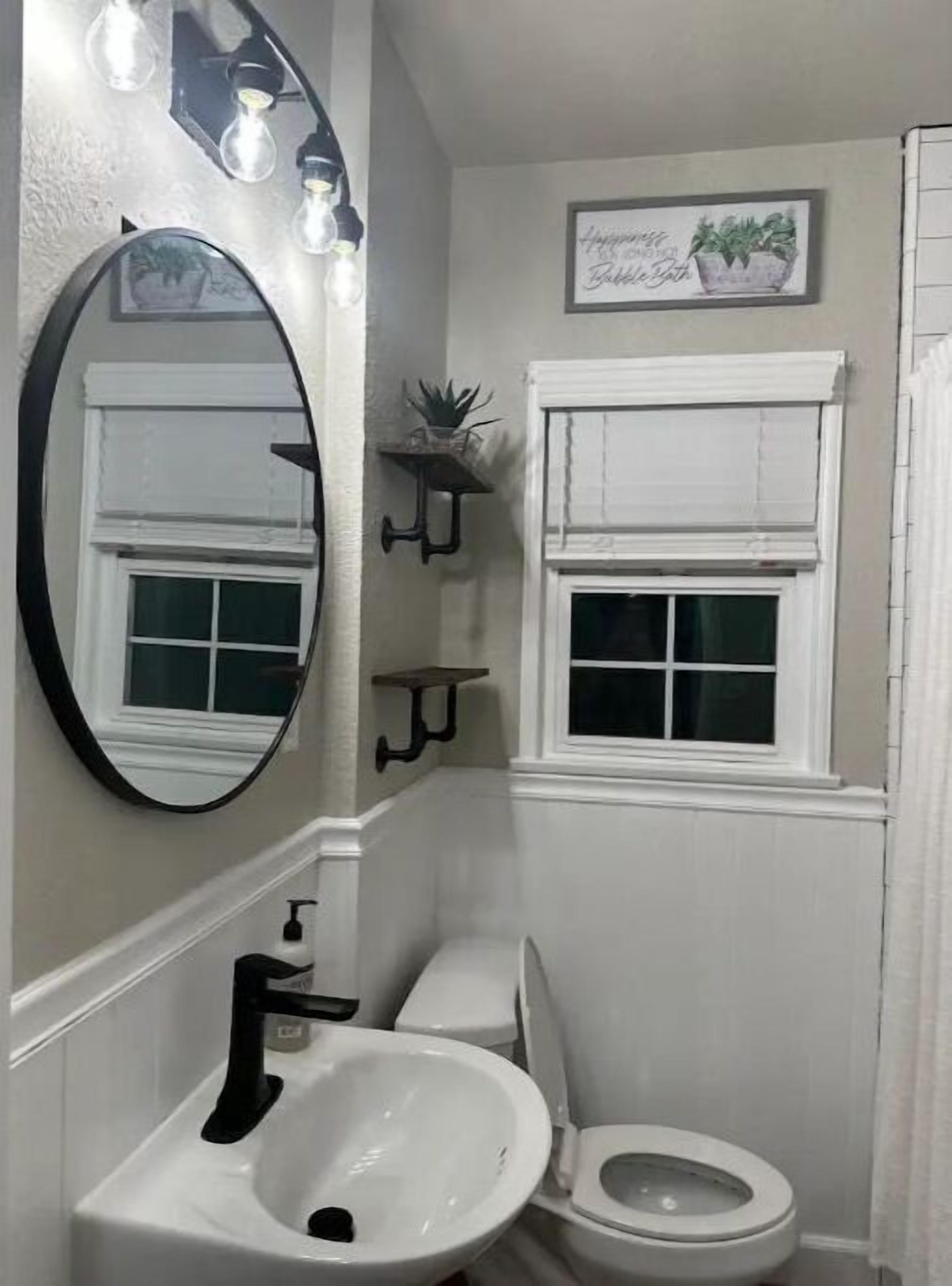 A bathroom with a white sink, toilet, and a mirror with lights. There are two wall-mounted shelves with plants and a framed picture above the toilet.