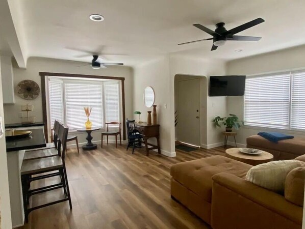 Un salon avec un canapé marron, une table à manger avec des chaises, un ventilateur de plafond et un téléviseur fixé au mur.