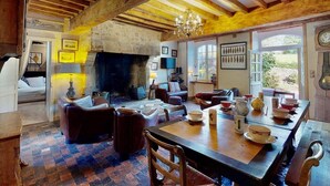 Une pièce rustique avec des poutres apparentes, une cheminée, une table à manger dressée et une vue sur l’extérieur.