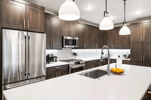A modern kitchen with dark wooden cabinets, stainless steel appliances, a white countertop, and pendant lighting.