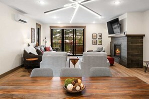 A modern living room with a fireplace, a dining table with a centerpiece, and a large window offering a view of the outdoors.