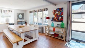 Uma área de refeições com uma mesa de madeira e bancos, uma janela com cortinas, um radiador e um quadro colorido na parede.