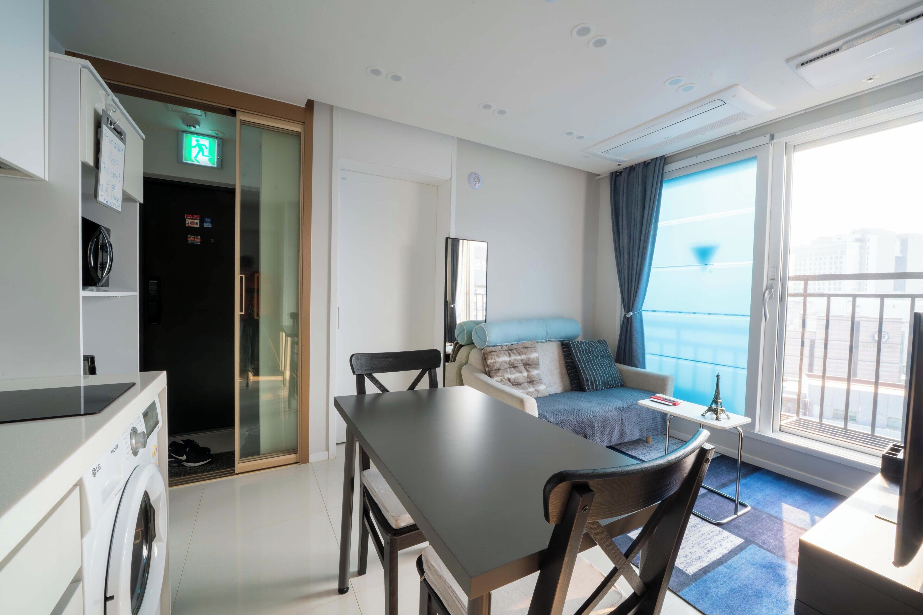 A hotel room with a dining area, a bed, a small table with a lamp, and a view of the city through the window.