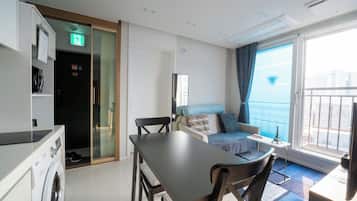 A hotel room with a dining area, a bed, a small table with a lamp, and a view of the city through the window.