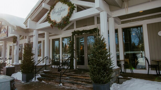 Entrada de un hotel con un letrero que dice "Persephone", con un amplio porche y escalones que conducen a la puerta, flanqueado por dos árboles de Navidad en macetas.
