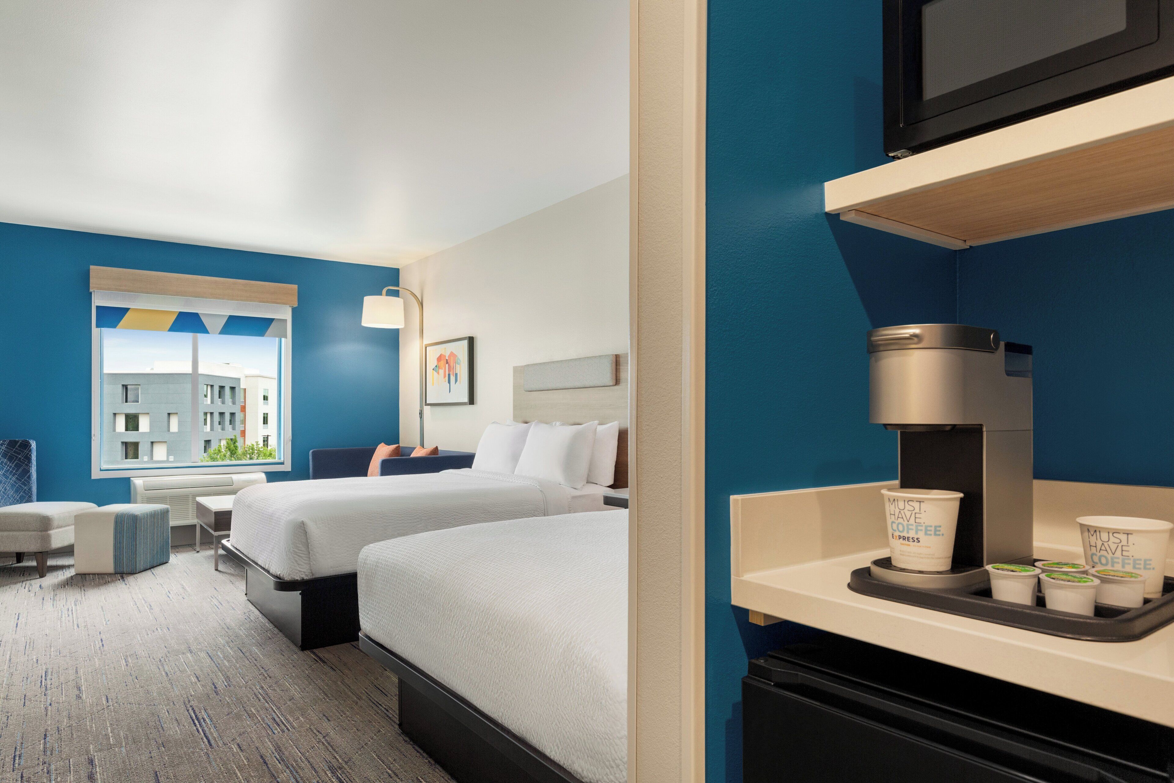 A hotel room with two beds, a desk, a chair, a coffee machine, and a window with a city view.