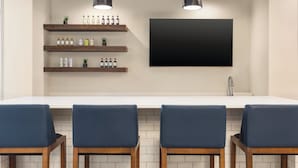 Una cocina moderna con encimera blanca, cuatro sillas azules, estantes de madera con botellas y un televisor negro grande montado en la pared.