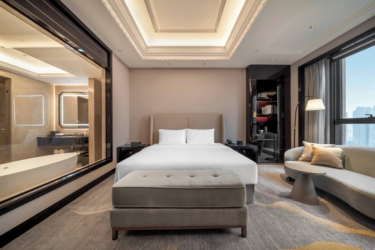 Ein modernes Hotelzimmer mit einem großen Bett, einem Sofa, einem kleinen Tisch und einem großen Fenster mit Blick auf die Stadt.