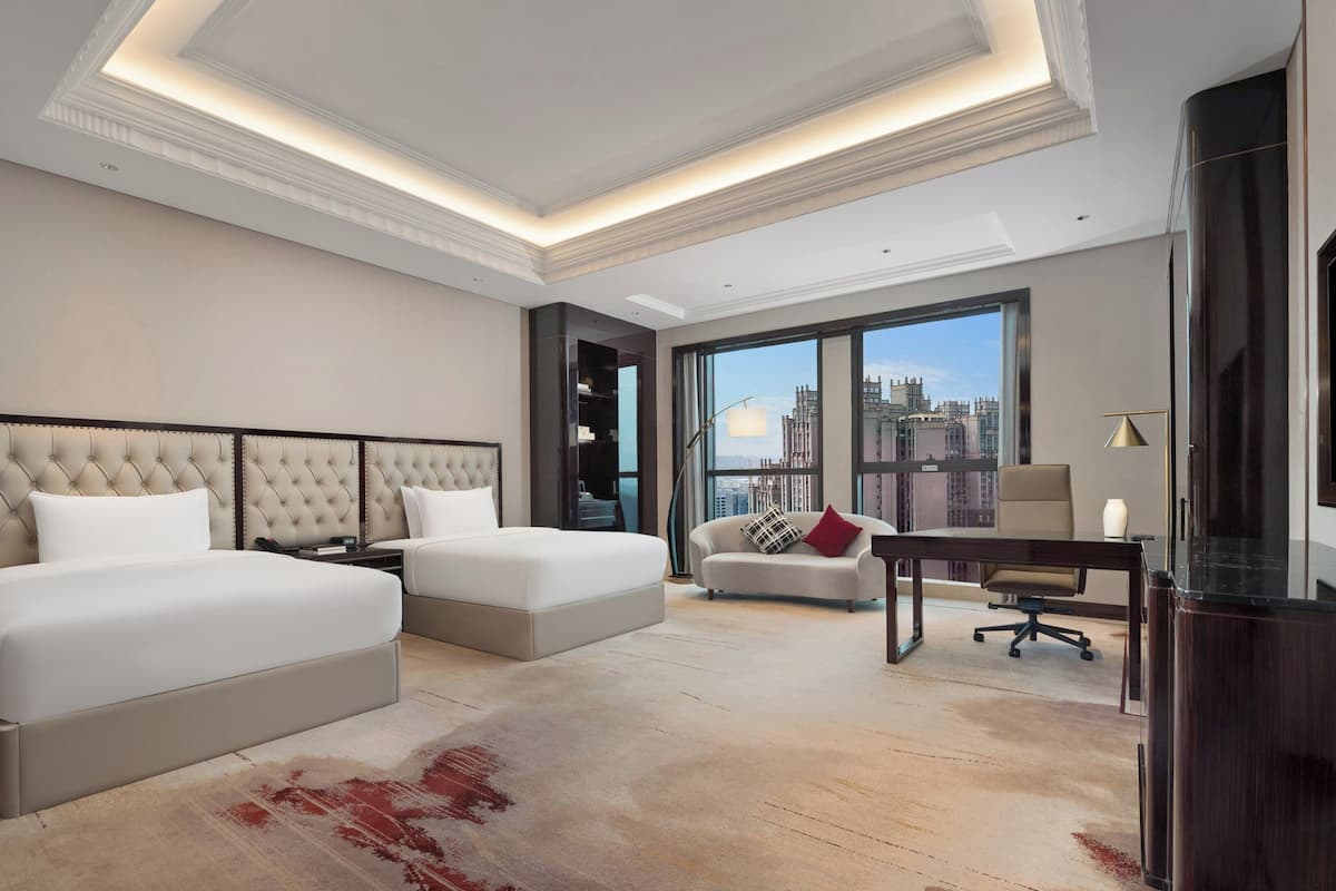 Ein Hotelzimmer mit zwei Betten, einem Schreibtisch, einem Sessel, einem Fernseher und einem großen Fenster mit Blick auf die Stadt.