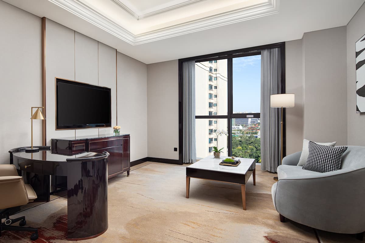 Ein modernes Hotelzimmer mit großem Fernseher, Schreibtisch, Sofa und Blick auf die Stadt durch hohe Fenster.