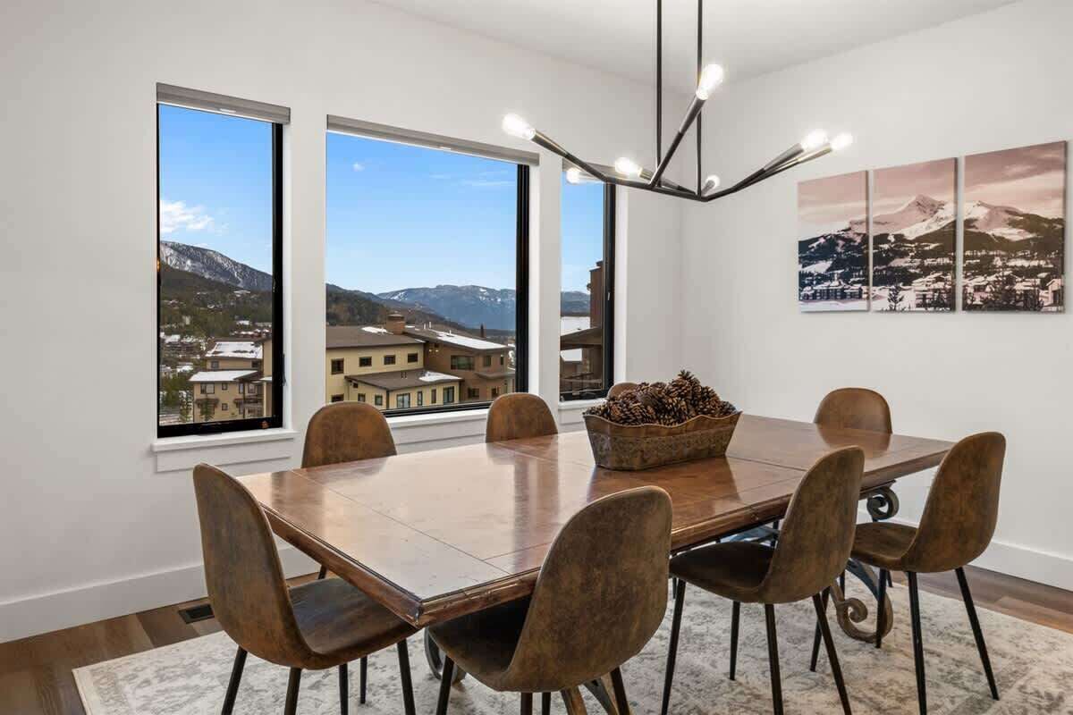 A dining room with a large wooden table, six chairs, and a chandelier. There are three windows offering a view of a mountainous landscape.