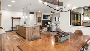 Una cocina moderna con armarios de madera, una isla central, iluminación colgante y un comedor con mesa y sillas de madera.