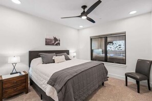 Ein Schlafzimmer mit Bett, Nachttisch, Stuhl, Deckenventilator und einem Fenster mit Blick auf eine verschneite Landschaft.