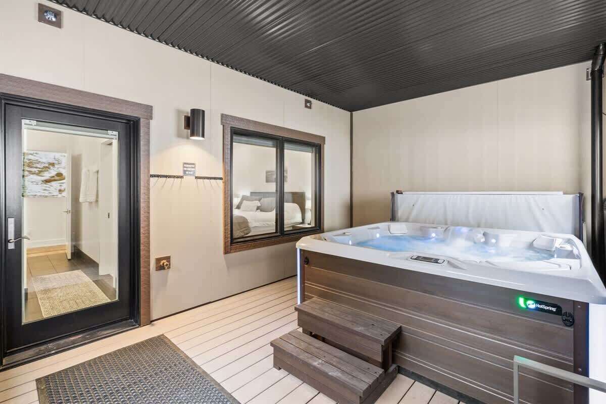 A modern spa room with a hot tub, wooden steps, a glass door, and a mirror.