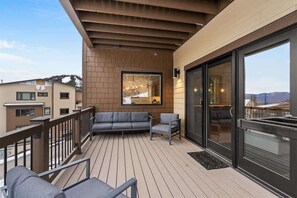 Een balkon met een houten terras, een grijze bank en stoelen, met uitzicht op een besneeuwde berg.