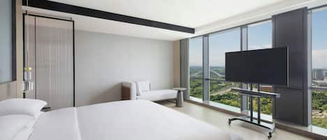 A modern hotel room with a large bed, a sofa, a TV on a stand, and a city view through large windows.