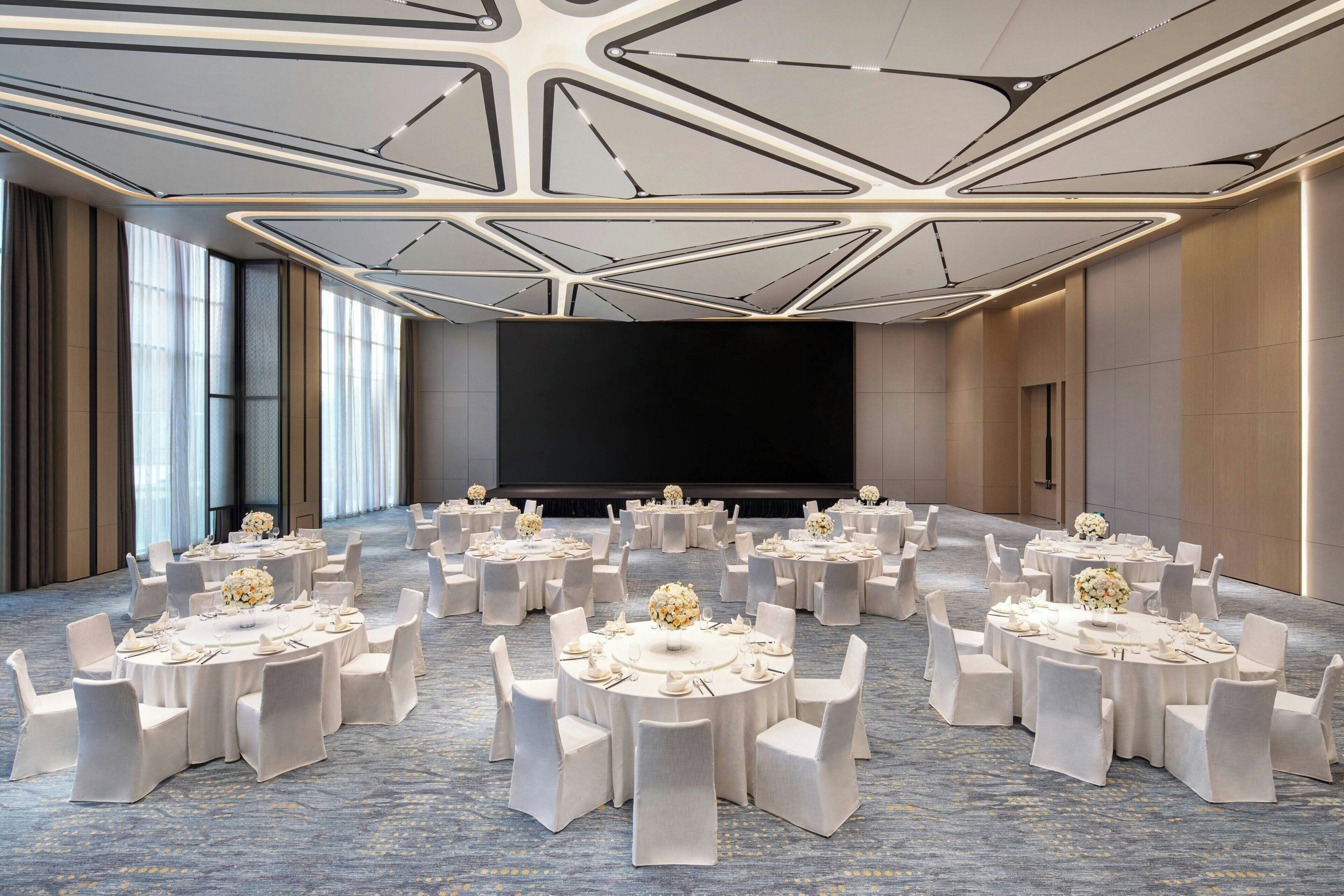 A conference room with multiple round tables, each set with white chairs and floral centerpieces. The room features a modern ceiling design with geometric patterns and large windows on one side.