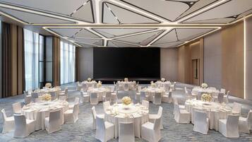 A conference room with multiple round tables, each set with white chairs and floral centerpieces. The room features a modern ceiling design with geometric patterns and large windows on one side.