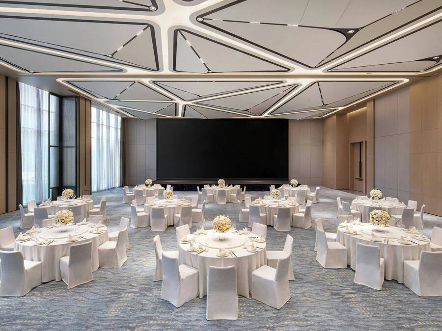 A conference room with multiple round tables, each set with white chairs and floral centerpieces. The room features a modern ceiling design with geometric patterns and large windows on one side.