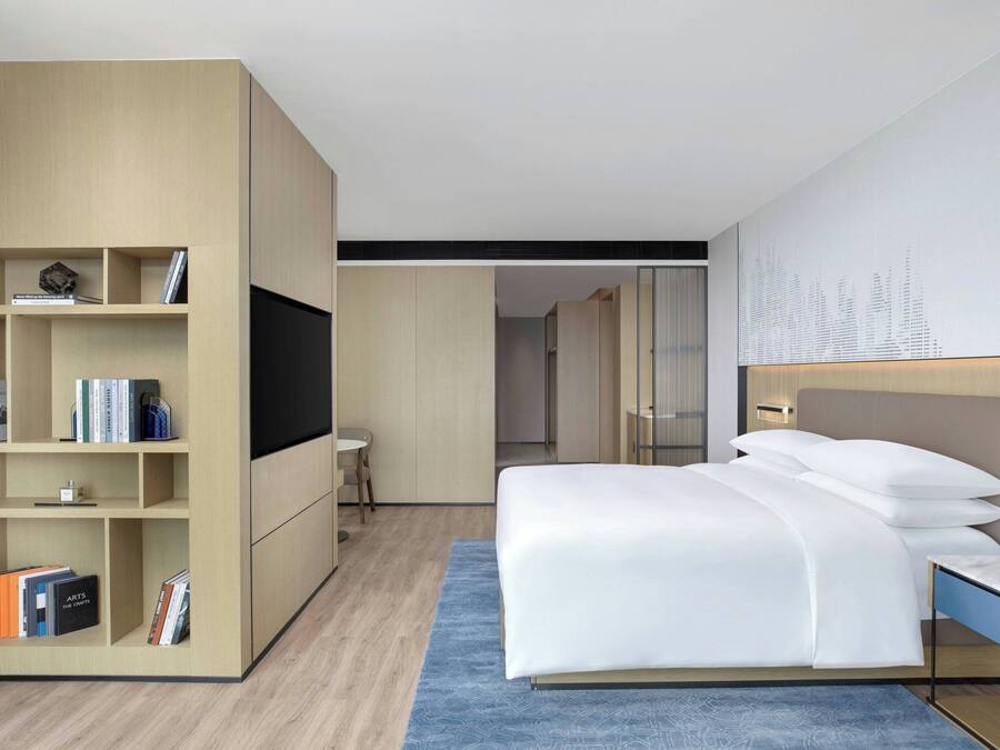 A modern hotel room with a large bed, a built-in bookshelf, a flat-screen TV, and a small desk.