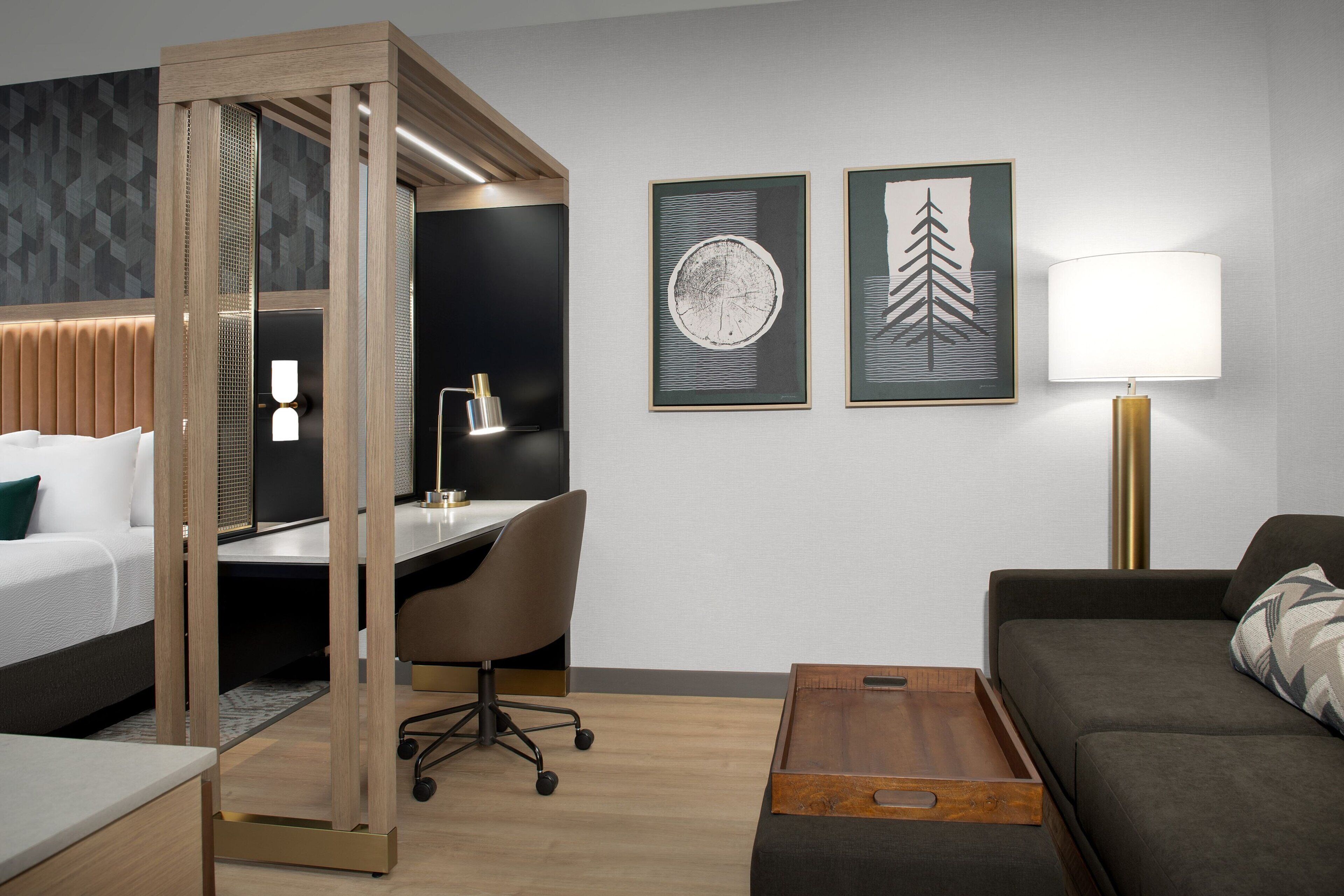 Una habitació d'hotel moderna amb llit, escriptori, cadira i sofà. Hi ha dues làmines amb marc a la paret i una làmpada al costat.