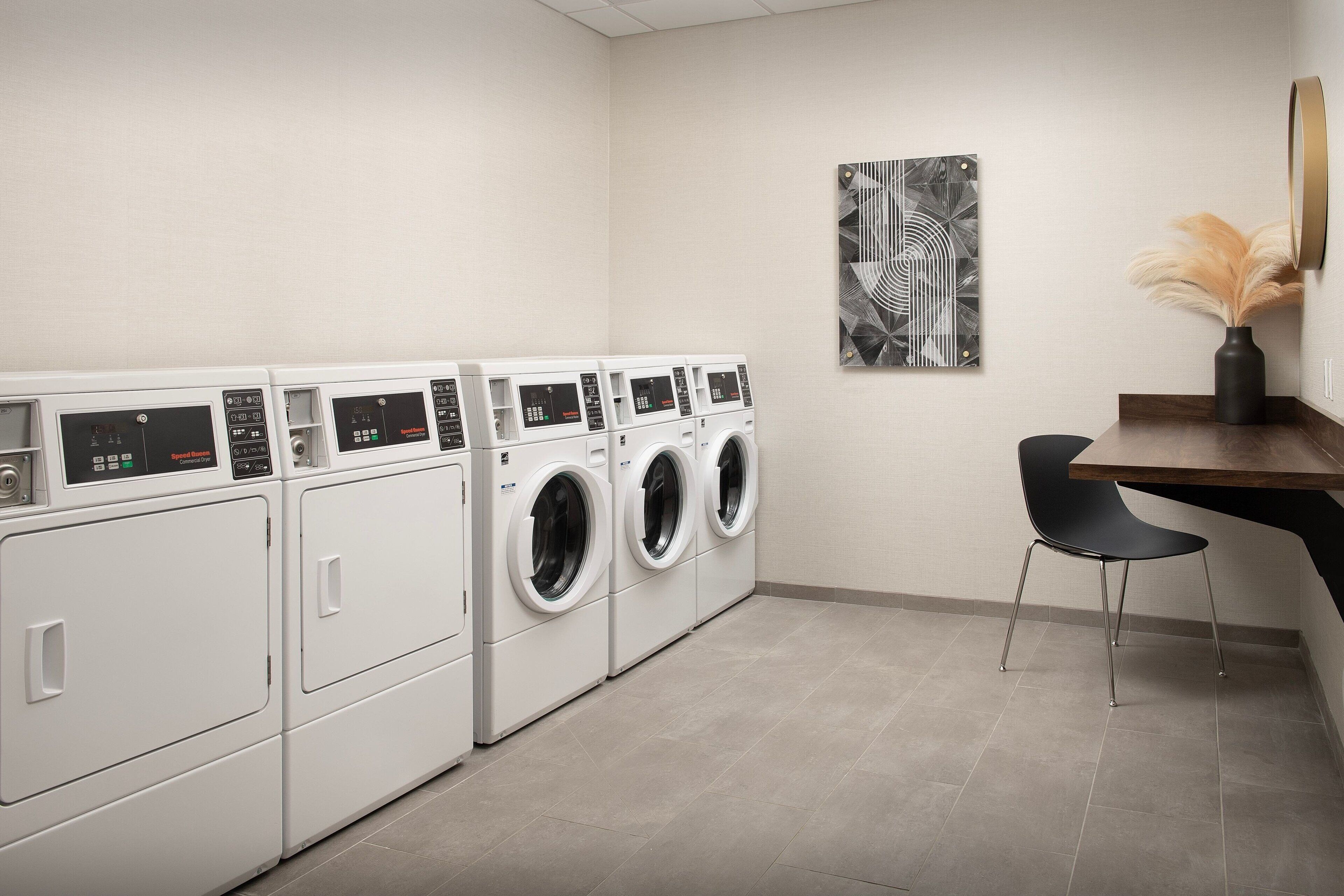 A laundry room with multiple washing machines, a small table, a chair, and a wall-mounted mirror.