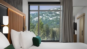 Ein Hotelzimmer mit großem Fenster und Blick auf die Berge, einem Bett mit weißer Bettwäsche und grünen Kissen sowie einem an der Wand montierten Fernseher.
