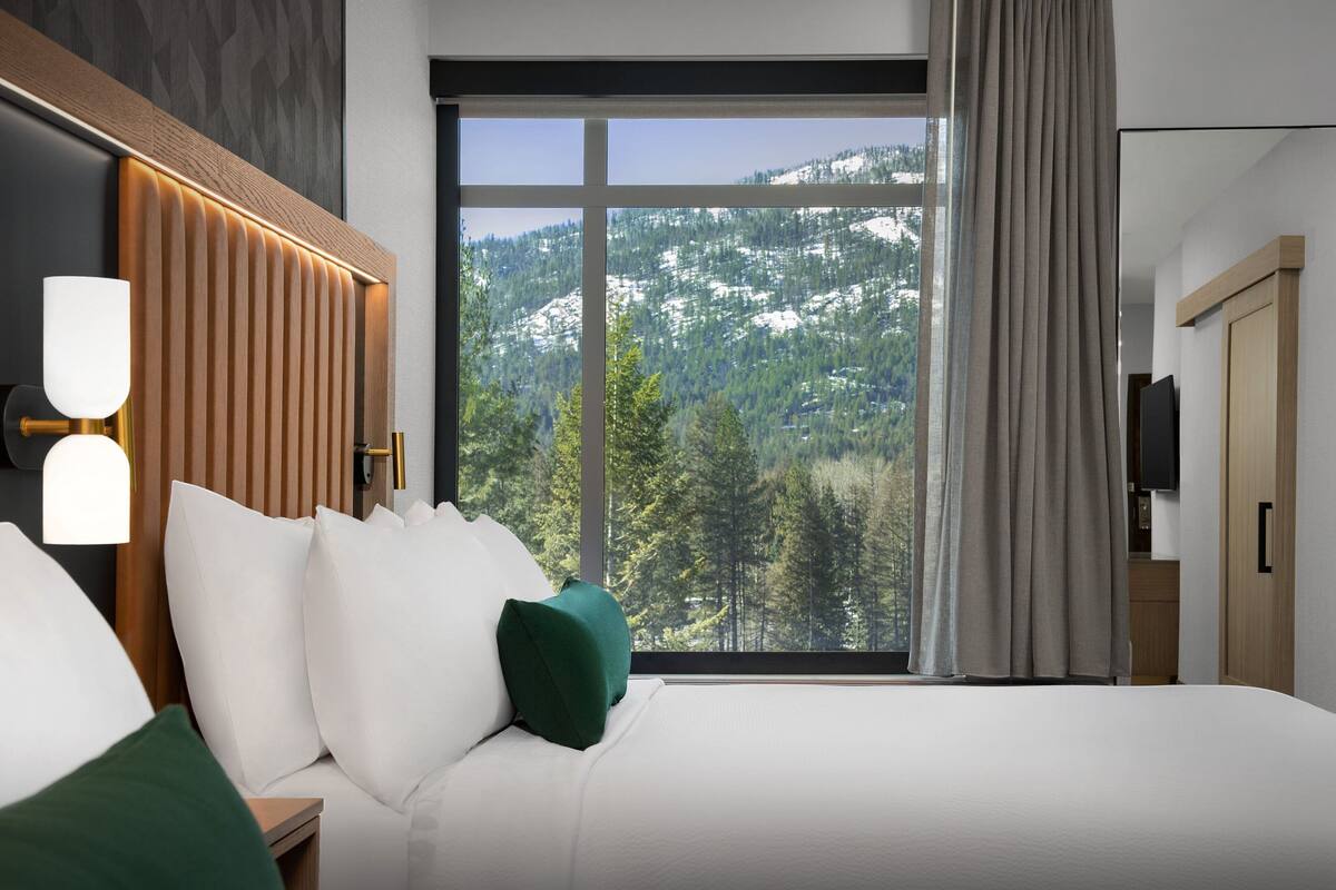 A hotel room with a large window offering a mountain view, a bed with white bedding and green pillows, and a wall-mounted TV.