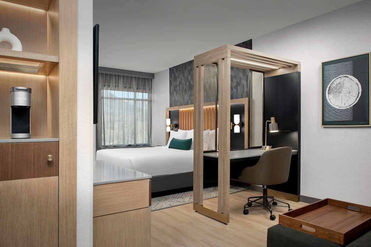 A modern hotel room with a bed, desk, chair, and a small wooden side table.