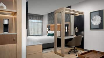 A modern hotel room with a bed, desk, chair, and a small wooden side table.