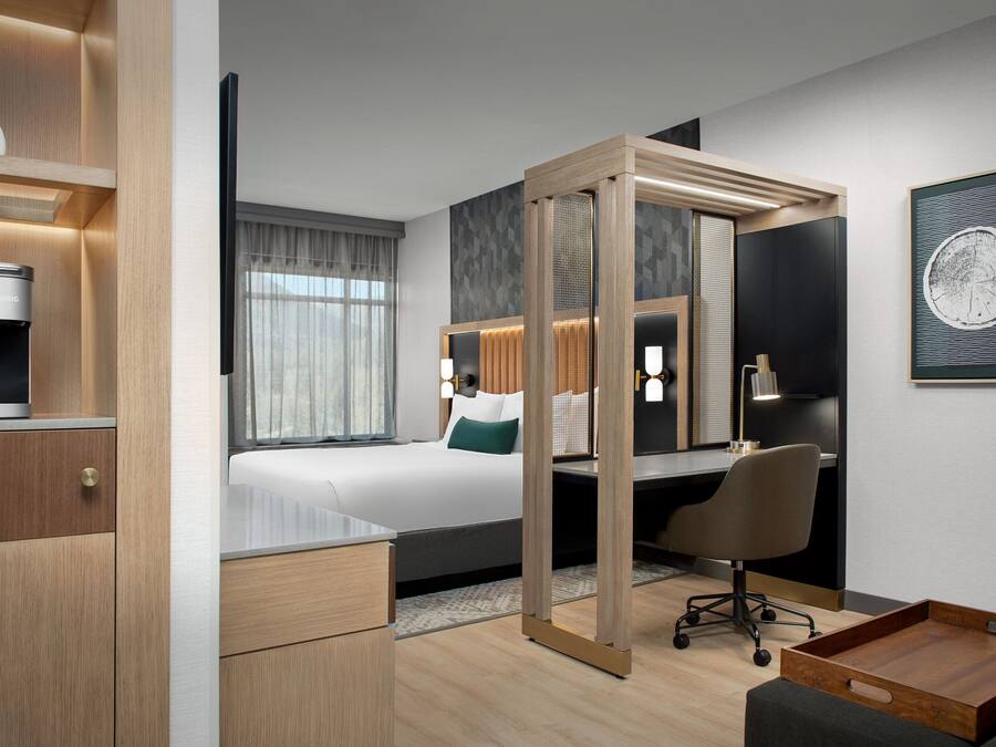 A modern hotel room with a bed, desk, chair, and a small wooden side table.