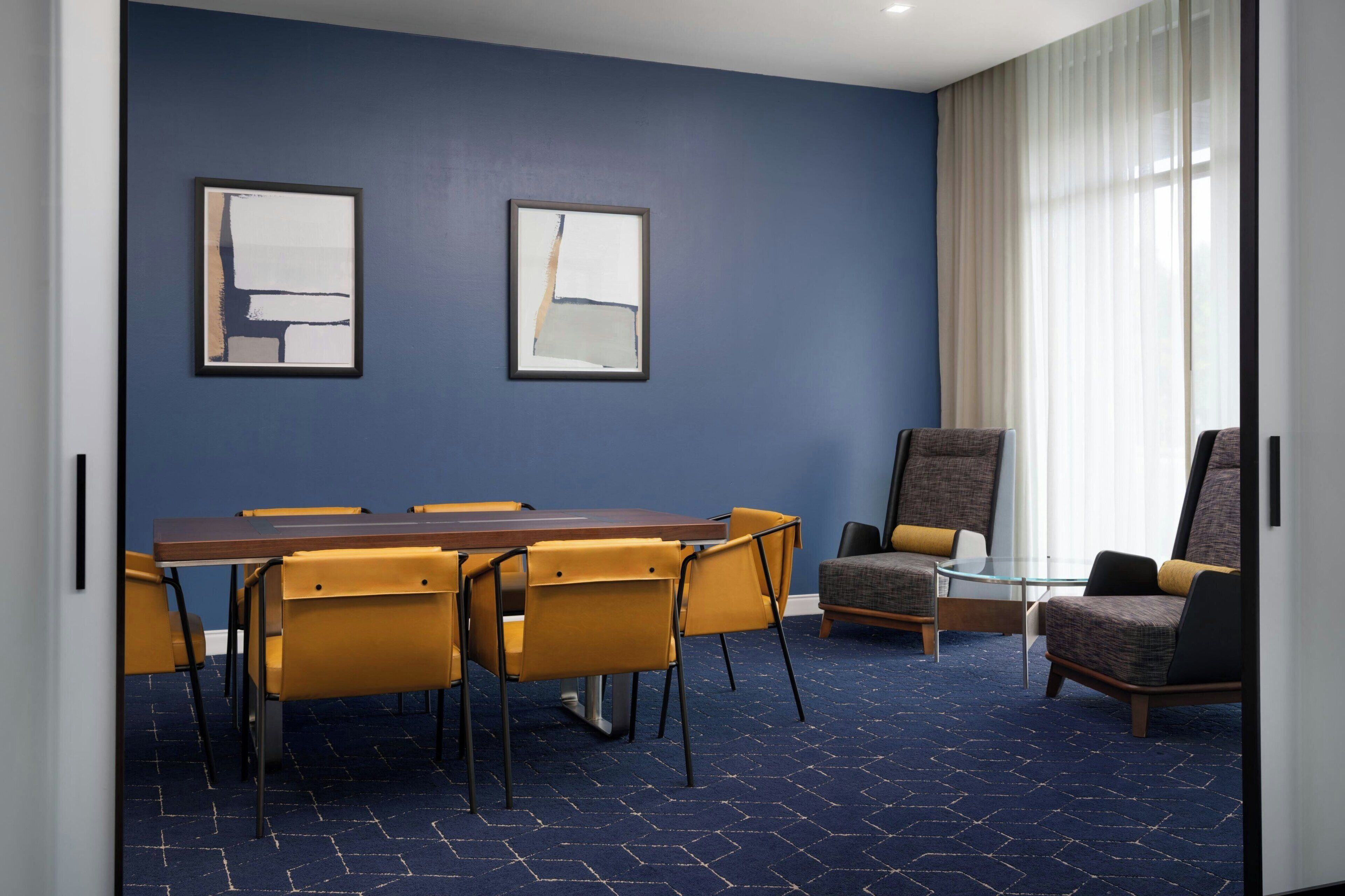 Une salle de conférence avec un mur bleu, une table en bois et des chaises jaunes. Deux tableaux abstraits encadrés sont accrochés au mur.