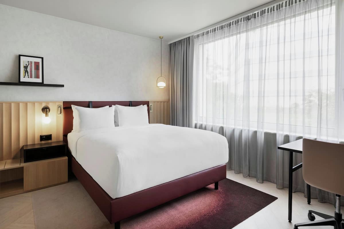 A modern hotel room with a large bed, bedside tables, a desk, and a window with sheer curtains.