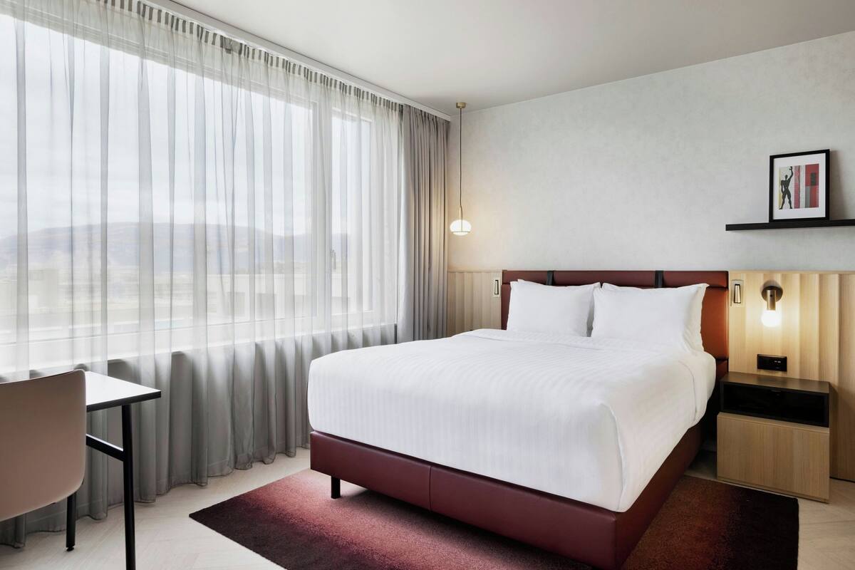 A hotel room with a large bed, a desk, and a chair. There is a window with curtains, a bedside table, and a wall-mounted light.
