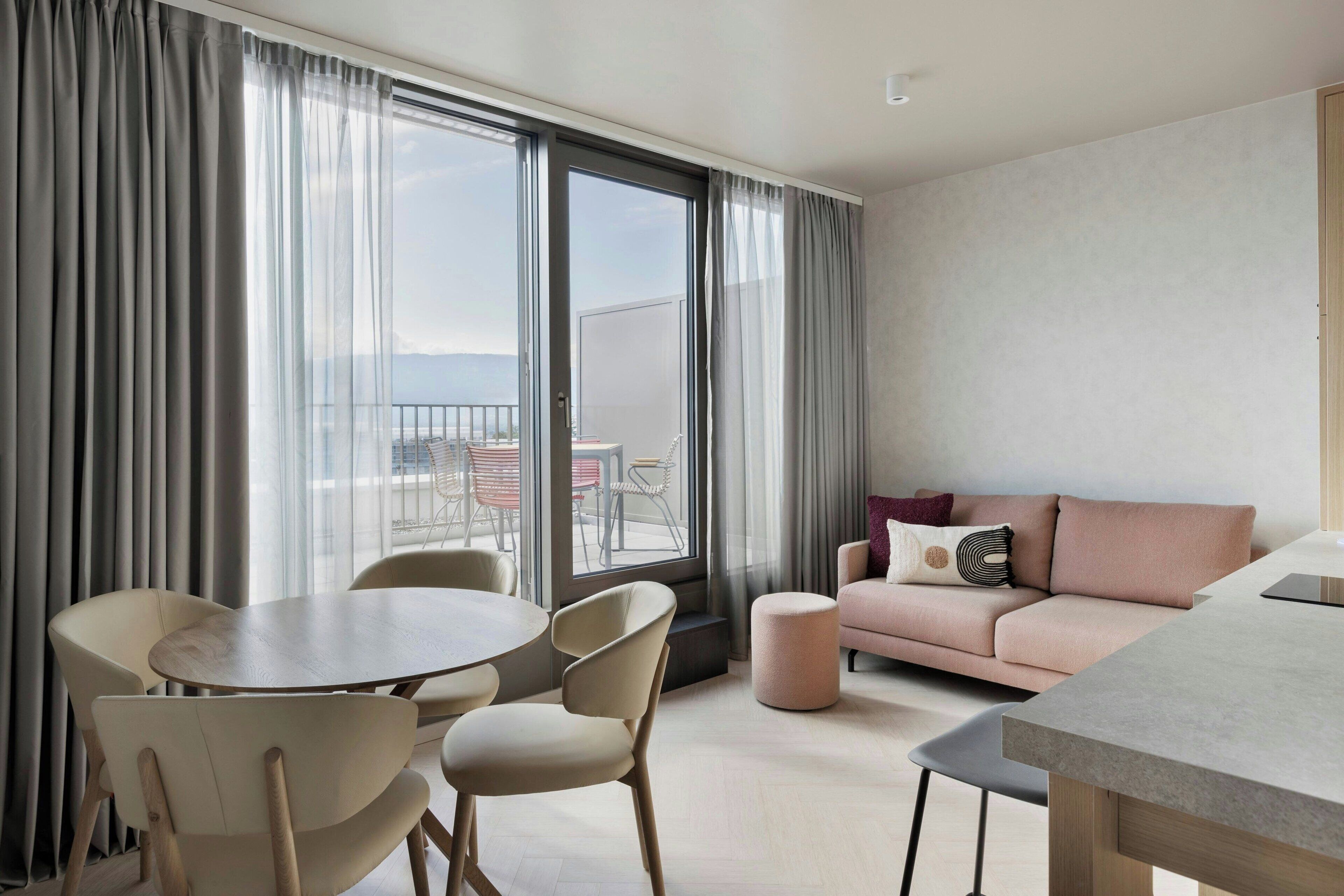 A modern hotel room with a dining area, a sofa, and a view of the city through large windows.