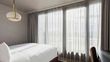 A hotel room with a large bed, a desk, and a view through sheer curtains.