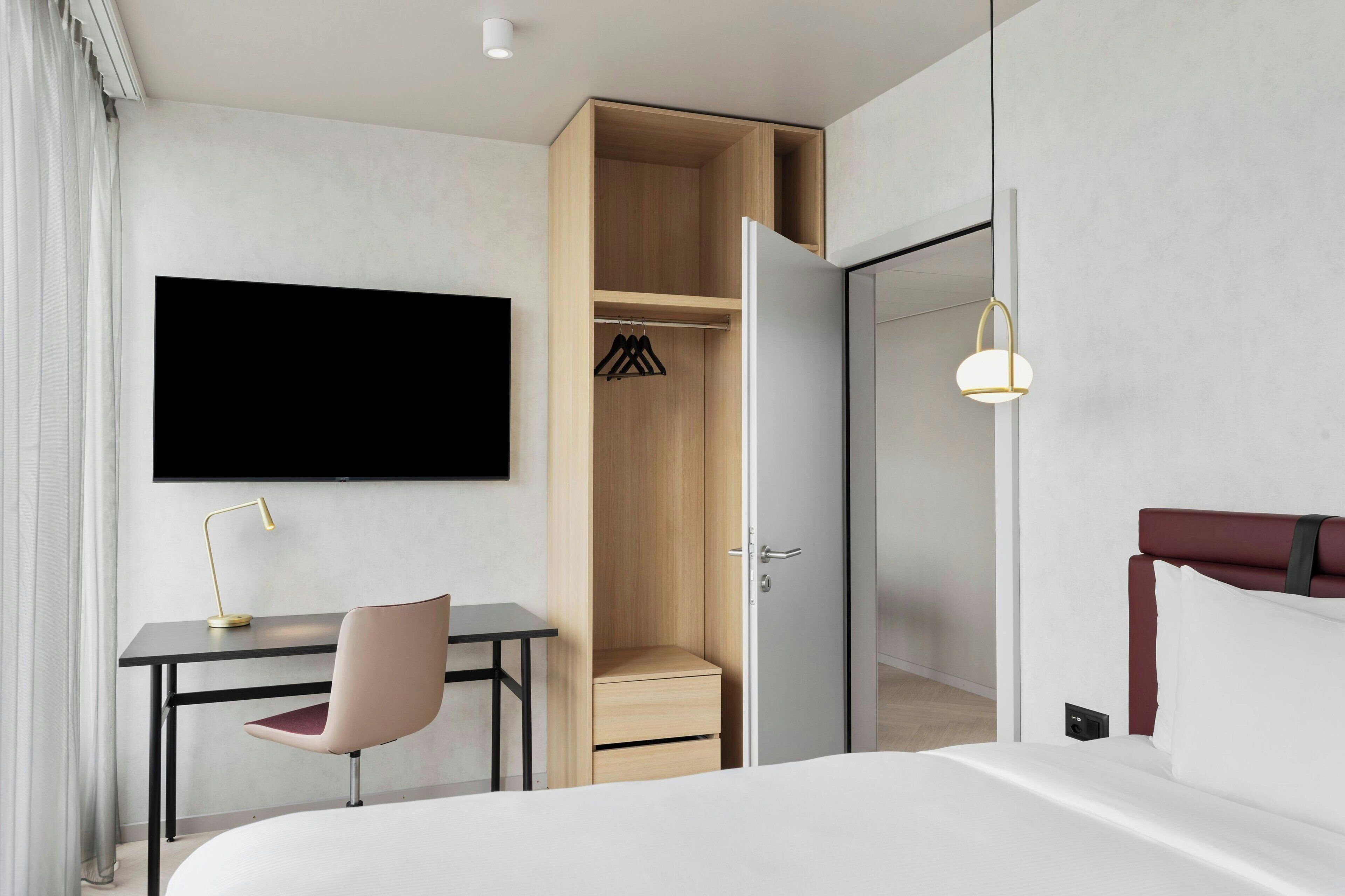 A modern hotel room with a bed, a desk, a chair, a TV mounted on the wall, and a wardrobe.