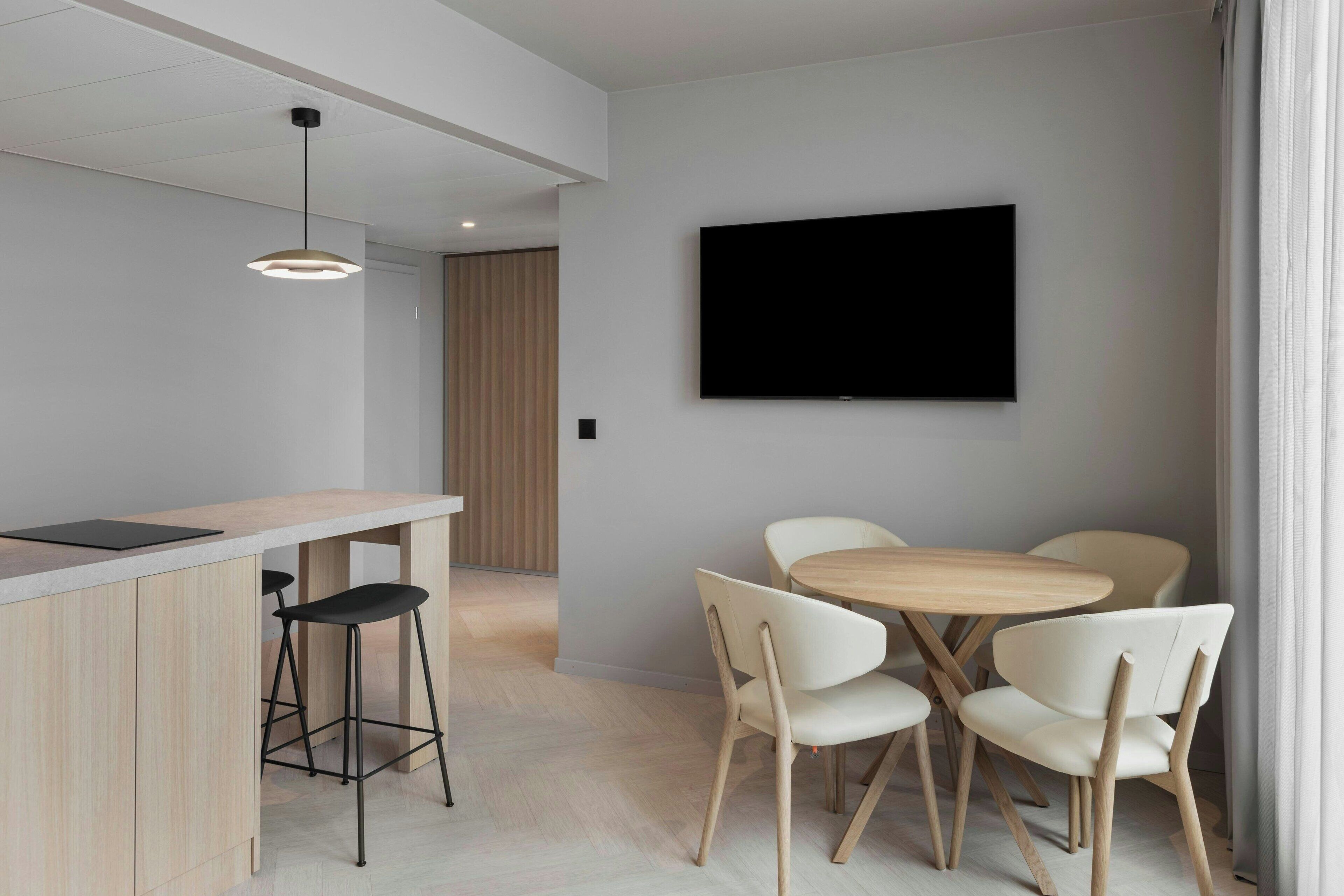 Una cocina moderna con encimera de madera y taburetes de bar, una zona de comedor con mesa redonda y sillas blancas, y un televisor de pantalla plana montado en la pared.