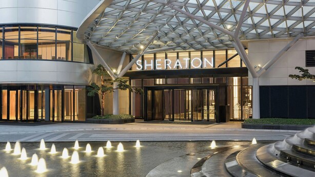 The Sheraton hotel entrance with a modern glass facade, a central atrium, and a water feature in front. - Sheraton Pujiang (Jinhua)