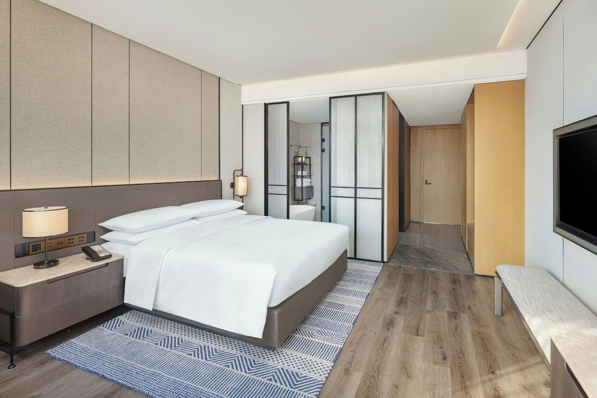 Ein modernes Hotelzimmer mit einem großen Bett, einem Nachttisch mit Lampe, einem Fernseher und einem durch eine Schiebetür einsehbarer Badezimmer.