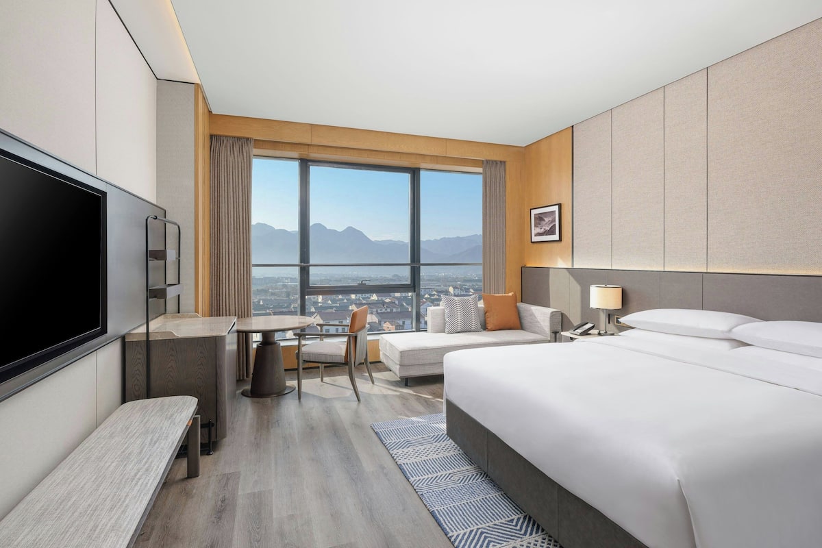 Ein modernes Hotelzimmer mit einem großen Bett, einem Schreibtisch, einem Sessel, einem Fernseher und Bergblick durch ein großes Fenster.