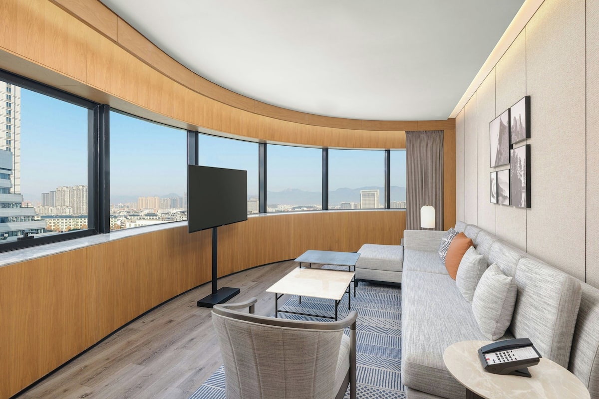 Ein modernes Wohnzimmer mit gewölbter Decke, großen Fenstern mit Blick auf die Stadt, einem Flachbildfernseher, einer Couch, einem Couchtisch und einem Telefon.