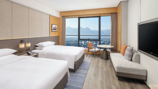 Habitación de hotel con dos camas, un sofá, un televisor, un escritorio y un ventanal grande con vista a la ciudad.