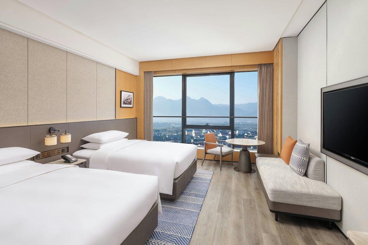 Ein Hotelzimmer mit zwei Betten, einem Sofa, einem Fernseher, einem Schreibtisch und einem großen Fenster mit Blick auf die Stadt.