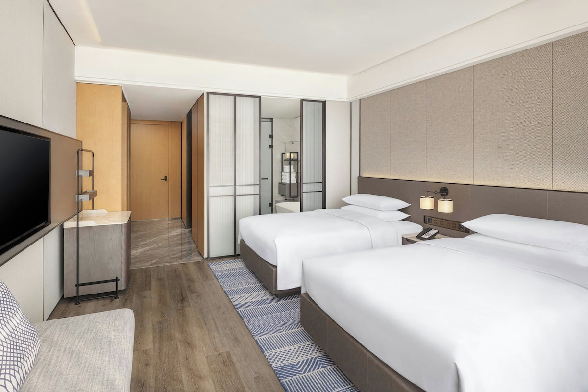 Ein Hotelzimmer mit zwei Betten, einem Fernseher, einem Schreibtisch und einem durch eine Schiebetür einsehbaren Badezimmer.